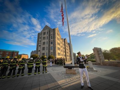 A cadet in dress uniform holds a rifle to shoot blank ammunition as first responders and cadets stand in the background ready to raise the American flag.